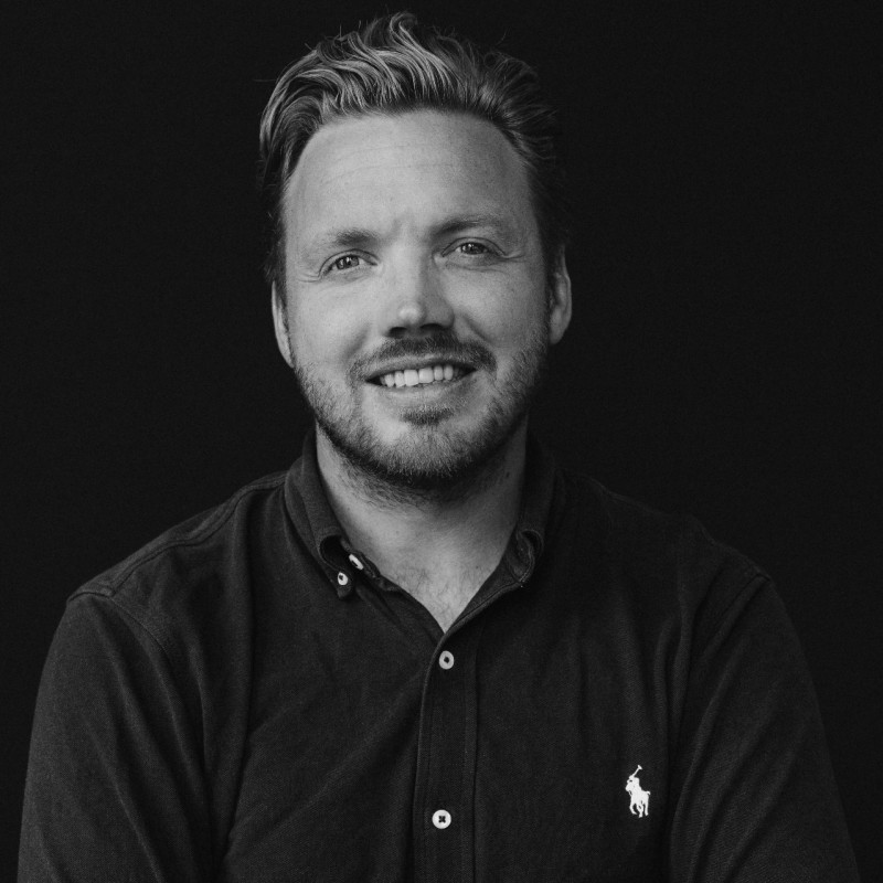 Portretfoto van een man met kort golvend haar en baard, glimlachend in een zwart overhemd met een klein paardlogo.